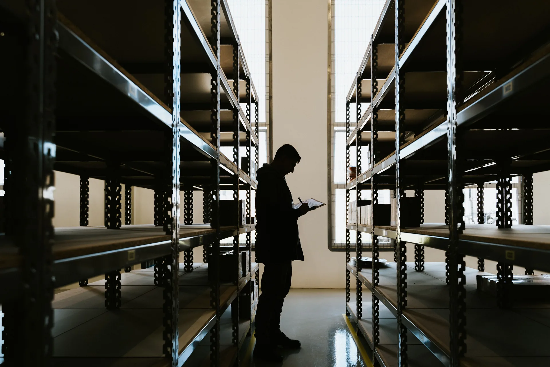 Silhouette of an inventory reviewer holding a clipboard in a warehouse aisle